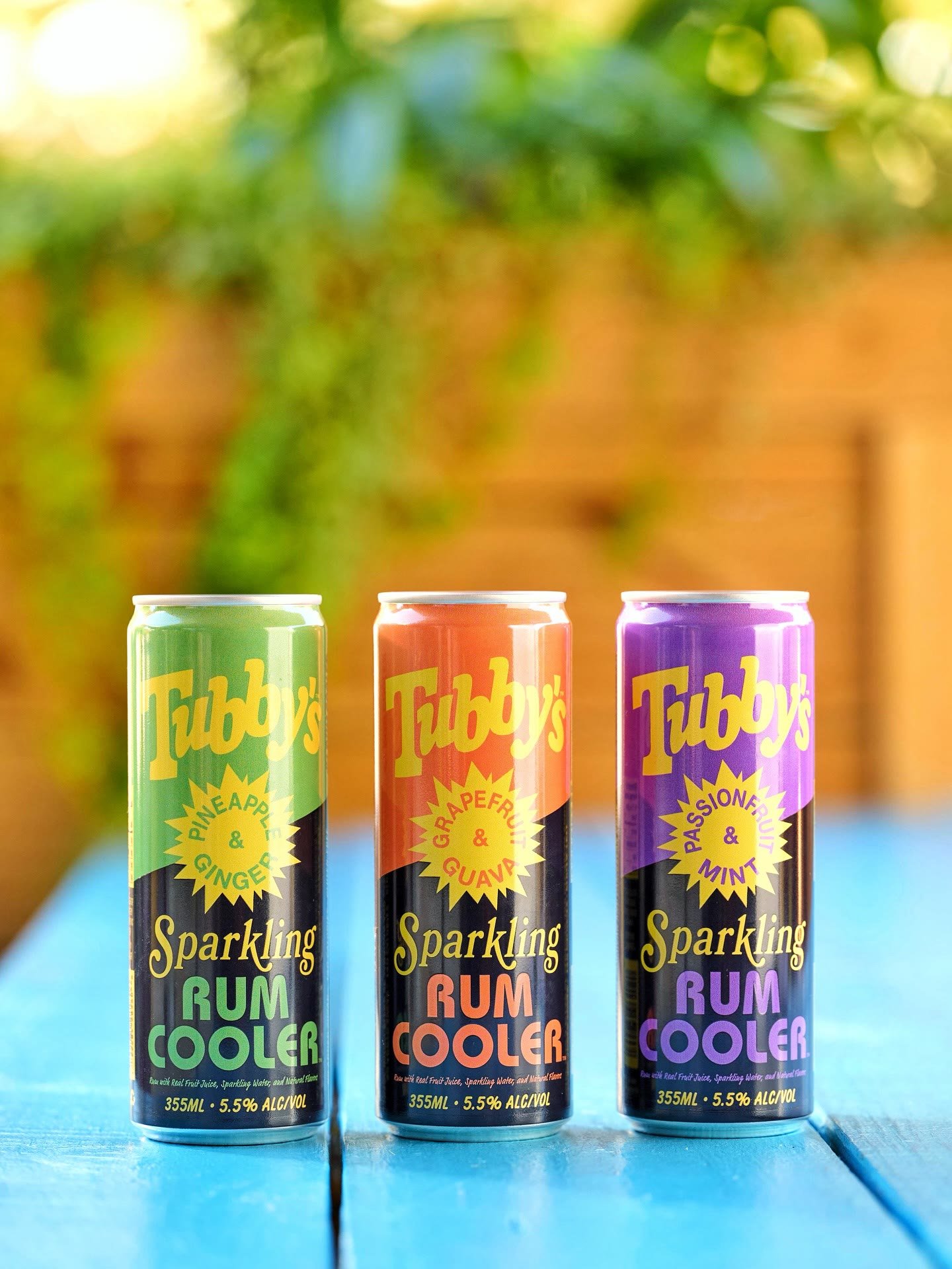 Three cans of Tubby's, Pineapple and Ginger, Grapefruit and Guava, and Passionfruit and Mint, on a turquoise table in the sun